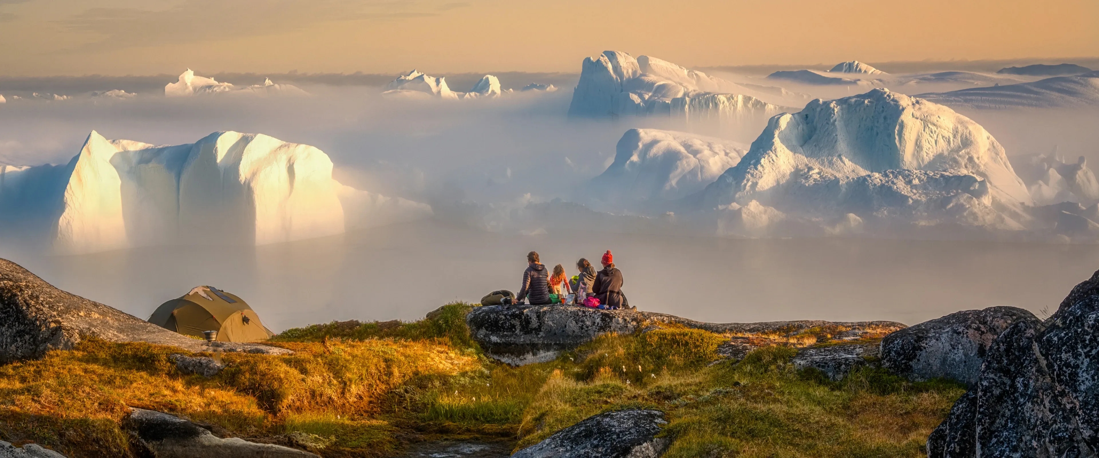 A group of people sit on a hill overlooking icebergs in Greenland